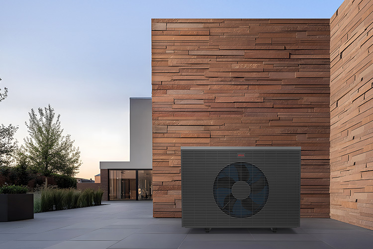Modern exterior view with a large, dark-coloured AEROTOP SPK heat pump in front of a tall, textured brick wall.