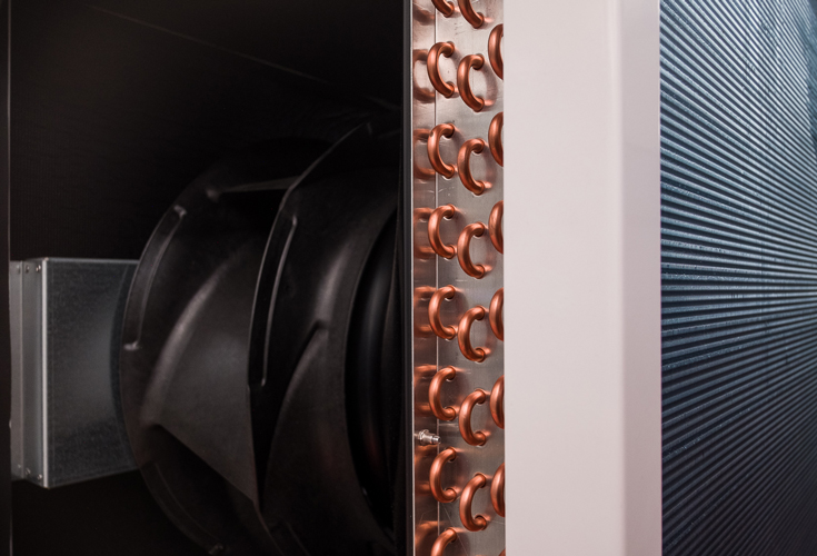 Close-up of a part of a heat generator, showing a black fan, copper pipes and a ribbed metal surface. 